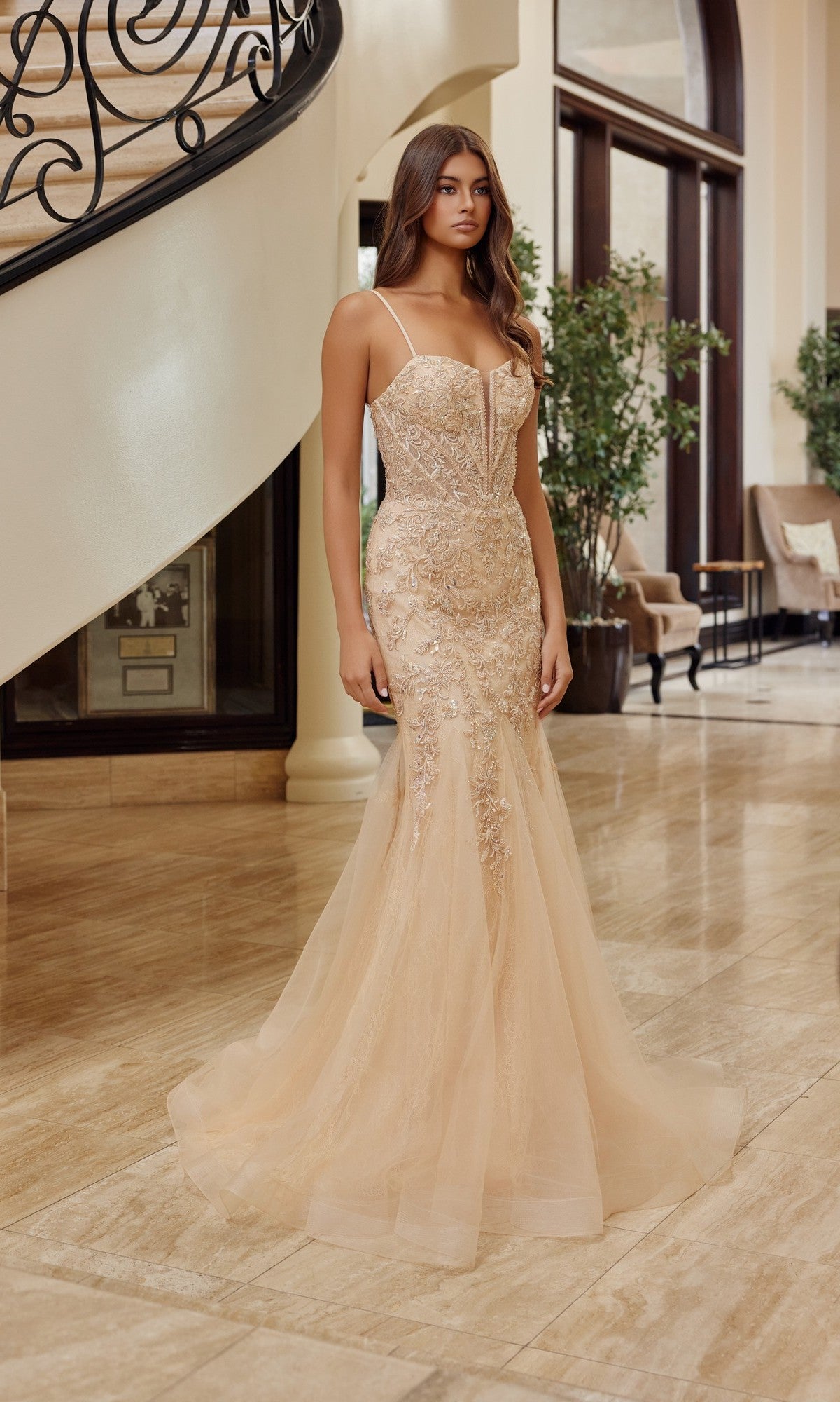 A woman stands indoors by a staircase with plants, wearing the Nox Anabel H1874 long prom dress in champagne. The fitted trumpet gown features lace details, thin straps, and delicate beaded sequin embroidery.